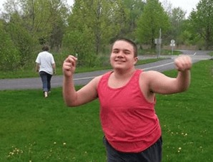 Photograph of a young man with black hair running and smiling. He is wearing a red tank top. Grass and trees are in the background.