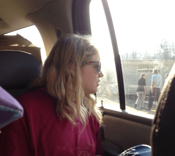 Photograph of Issy Stapleton. Light skinned blonde girl with shoulder length hair looks out a car window. She is wearing sunglasses and a pink tshirt.
