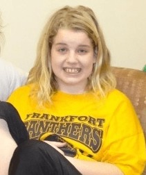 Photograph of a blonde girl smiling at the camera. She is wearing a yellow tshirt.
