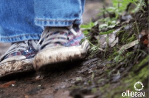 Photograph of lower part of jeans and multicolored plaid sneakers. The sneakers have mud on them and the person wearing them is standing on damp earth . There is green moss and foliage on the right side of the image and Ollibean logo in white. Ollibean logo is a circle composed of various shapes and sizes of equal signs and the word Ollibean.
