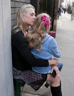 Photograph of blonde mother holding her blonde daughter in her lap.