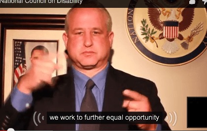 Image description: photograph of light skinned man with light hair using sign language . He is wearing a black blazer, blue shirt, and dark tie. Captioned white text on a black background reads" we work to further equal opportunity".